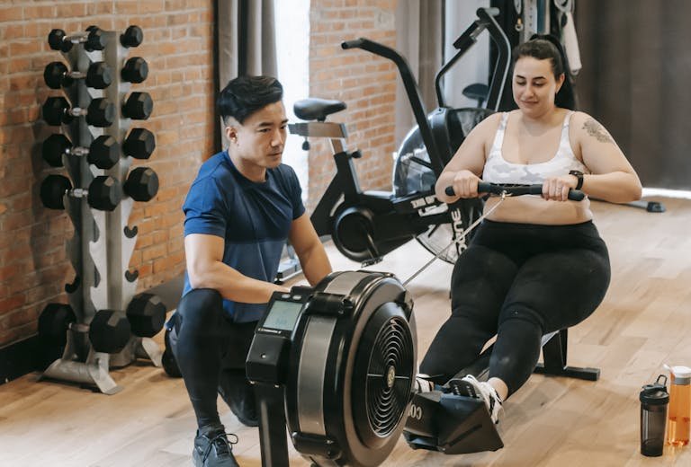 Positive young plus size female in leggings and crop top doing abs exercise on rowing machine during intense workout in modern gym with fit Asian male trainer