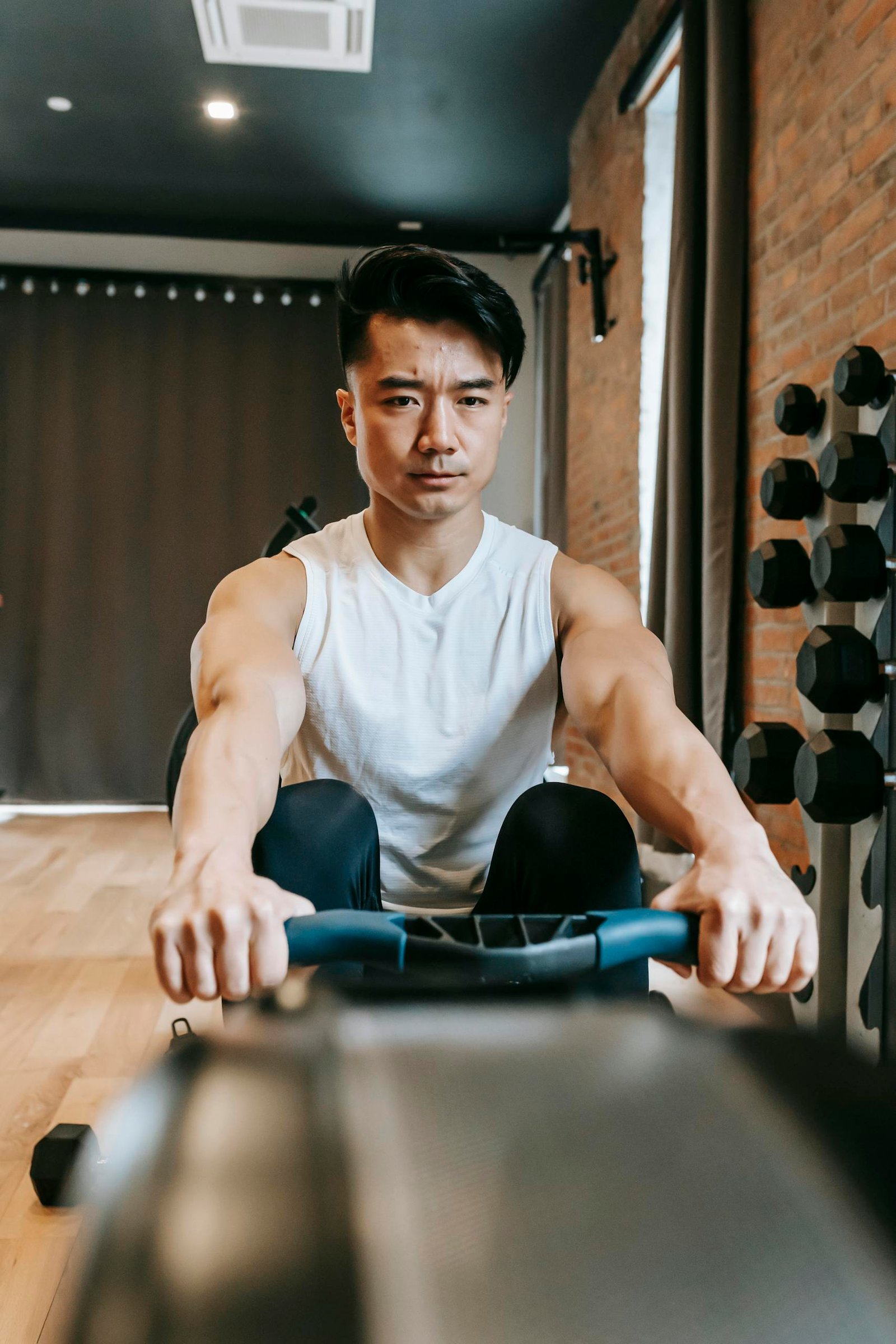 Fit ethnic sporty young focused male working out in contemporary gym with brick walls sitting on rowing machine