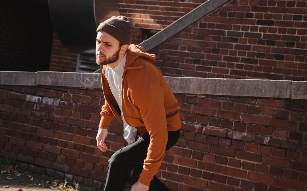 Serious energetic male tracer in hoodie and hat jumping from brick construction while practicing parkour on sunny day